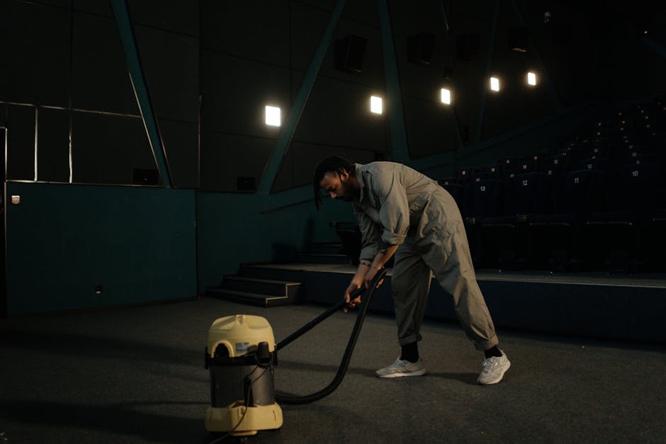 A person dressed in a light-colored jumpsuit is operating a portable vacuum cleaner on a carpeted floor in an indoor setting, likely a conference or presentation room with dark walls and a white proje