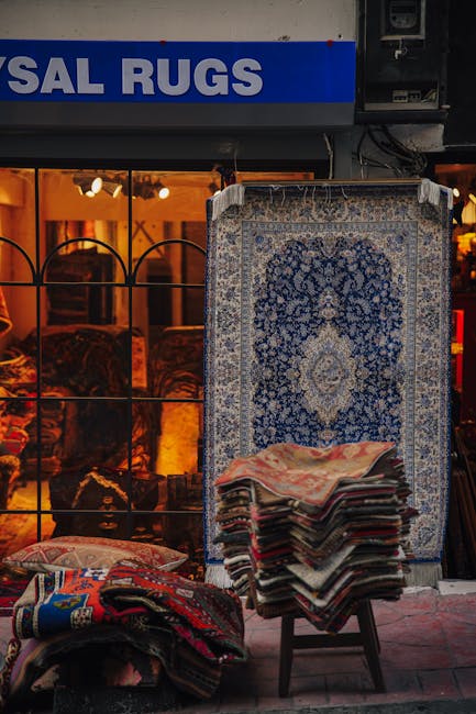 A row of colorful oriental rugs with intricate patterns and detailed designs hanging on a clothesline outdoors in a garden setting. The rugs feature shades of red, navy, beige, and gray, made of mater