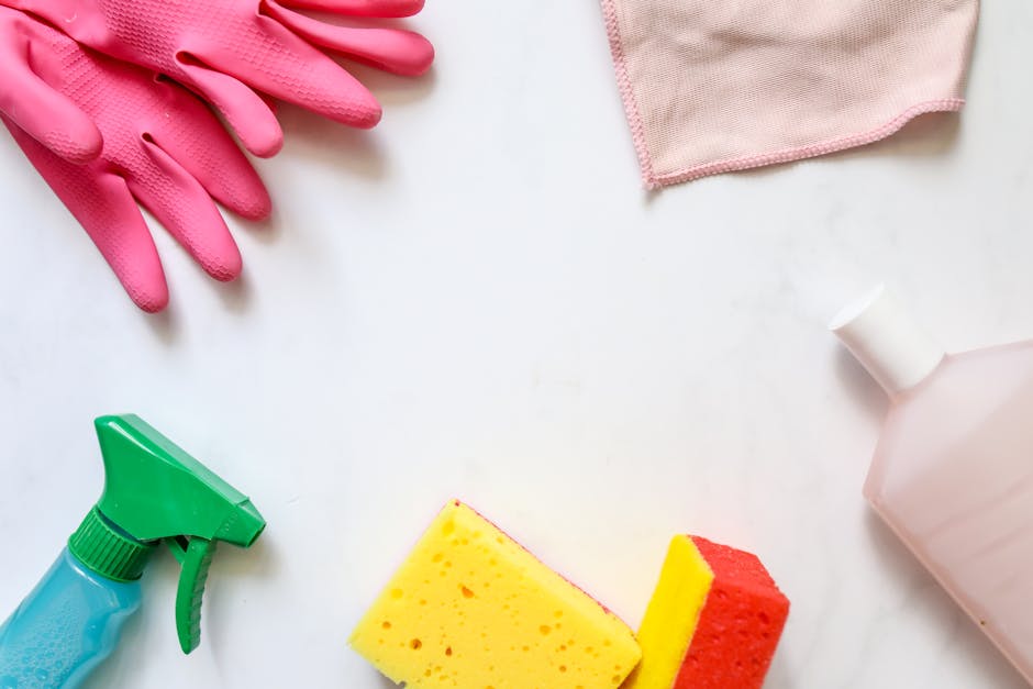 Arrangement of cleaning supplies on a white surface, including a pair of pink rubber gloves, a pink cleaning cloth, a green spray bottle with a blue base, a pink plastic bottle, and two kitchen sponge